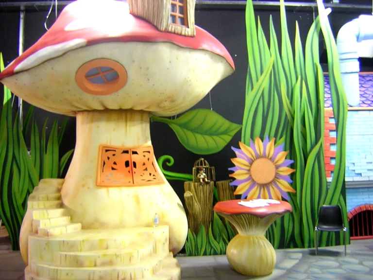 View of the fairy-tale set with a giant mushroom with red polka-dot cap, tall cartoon green grass painted on the walls, tropical leaves, an oversize daisy-flower on the right with orange and fuchsia petals, a small cage with a bird, a yellow spiral staircase on the left