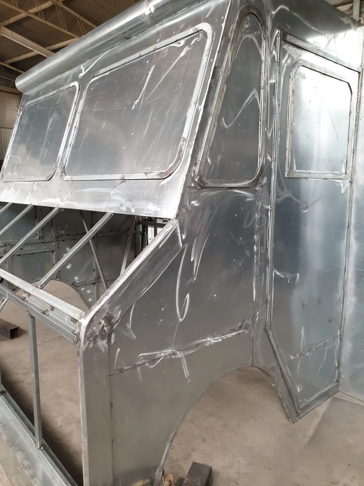 Raw welded sheet-metal body inside the Creation Art workshops: a metal van-truck silhouette, a cab with three rectangular windows in the walls, a square pre-paint structure, the industrial workshop floor visible