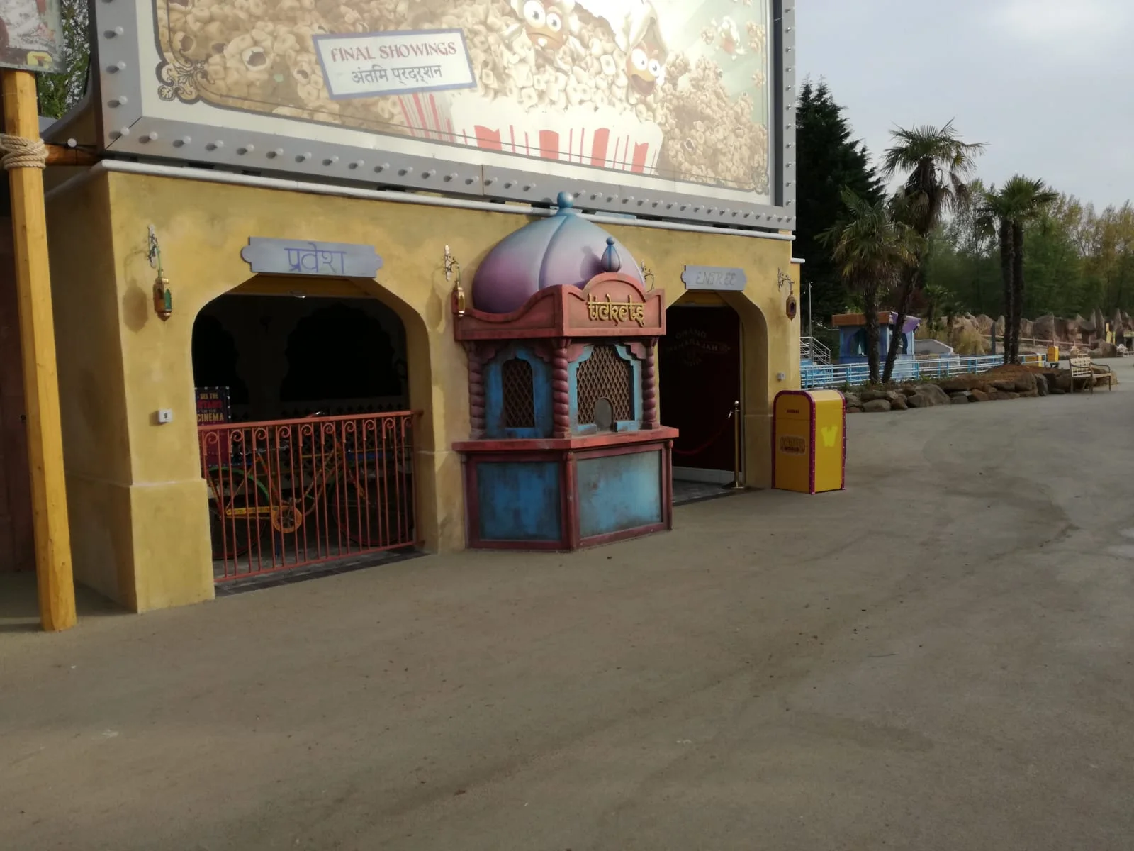 Mughal-style temple kiosk installed in the Bollywood area: a small violet-and-gold structure with a pink-magenta onion dome on top, a base in painted faux stone, the yellow walls of the building behind with a white 'FINAL SHOWINGS' sign, Hindu-temple-shaped entrance arches