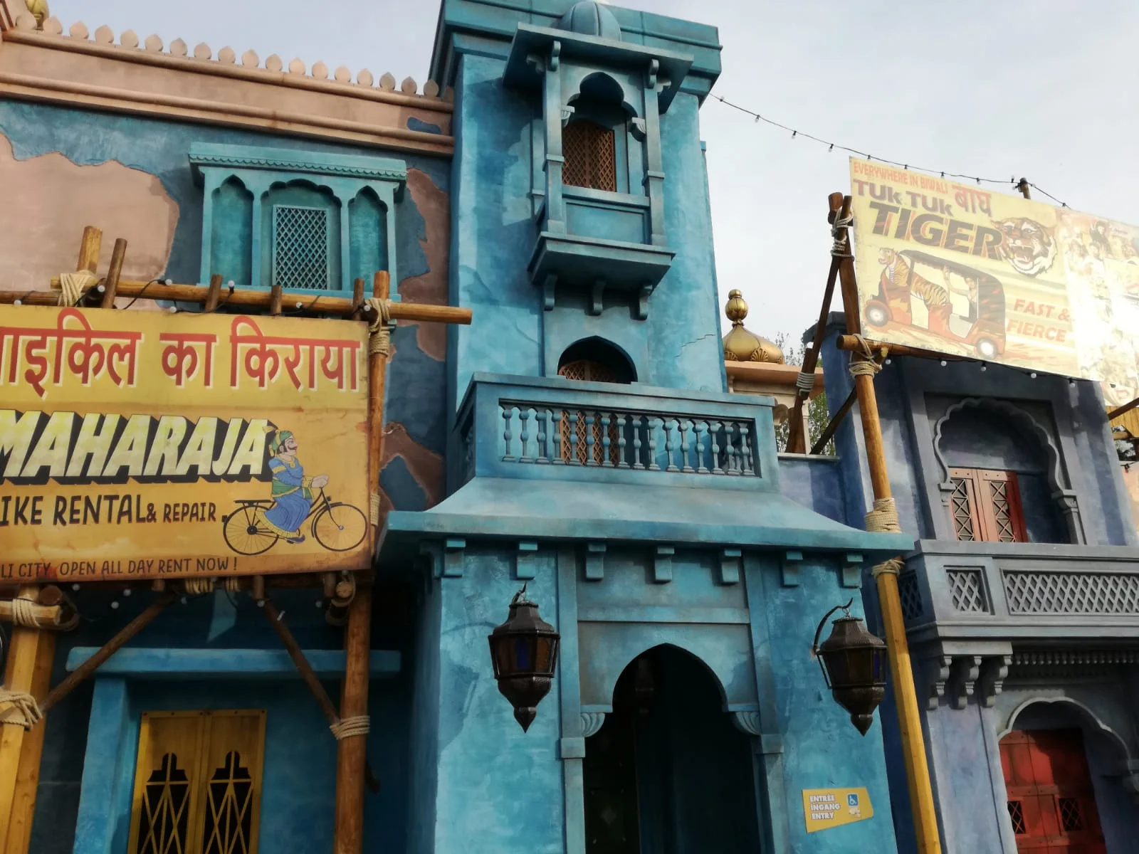 Two-storey scenographic buildings in Indo-Mughal style installed at Walibi: saturated turquoise-blue walls, balconies with white balustrades, lattice windows, a central tower with a gilded onion dome, hanging Arab lanterns; in the foreground posters reading 'MAHARAJA Bike Rental & Repair' on yellow and 'Tuk Tuk Tiger Fast & Fierce' on red with a tiger