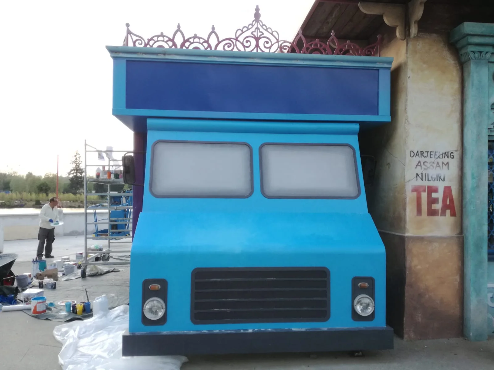 Front view of the finished blue food truck: a metallic-blue cab with two square windows, a large windscreen, round headlamps, a black grille, a front decoration of orange and gold flowers forming a kind of mandala; above it the pink-magenta openwork Mughal crown; an outdoor build site with a ladder and Ivano in the distance
