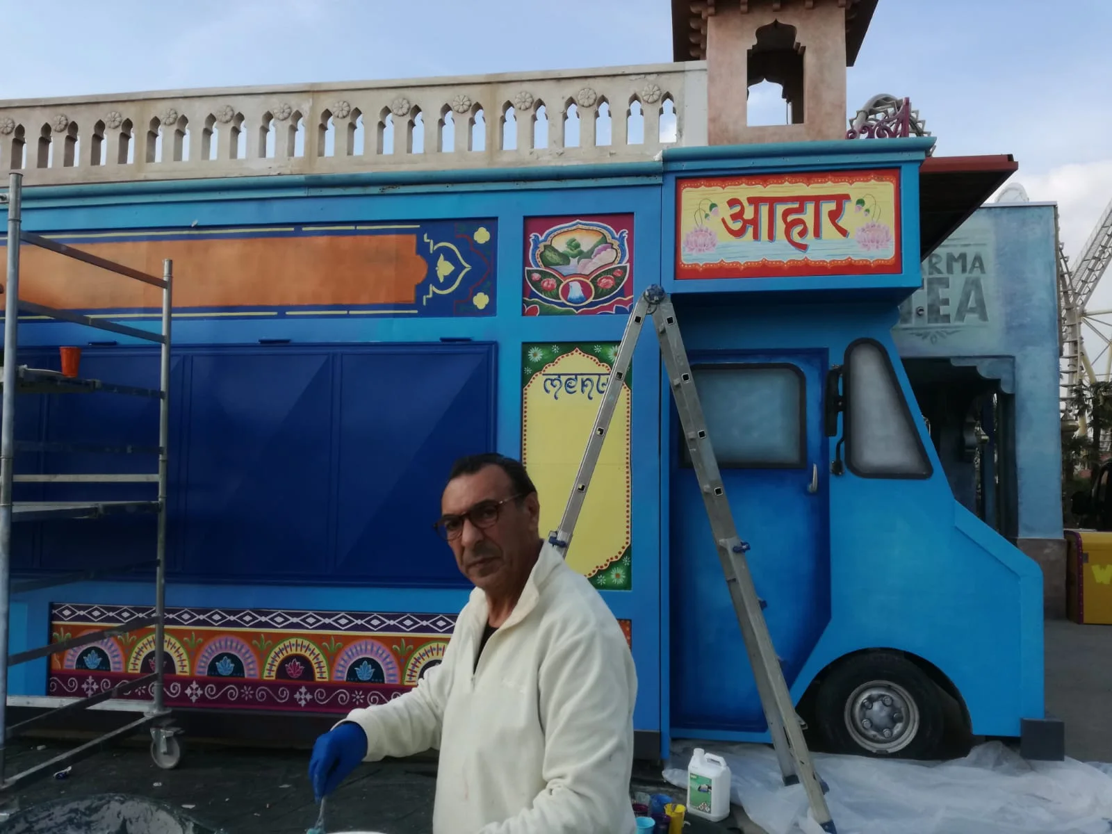 Ivano Ferrario, owner of Creation Art, on site at Walibi in the Bollywood area: white coat, glasses, blue glove, painting on the blue Shiva food truck decorated with Indian friezes; in the background the build-site ladder, the kiosk with the Hindi sign आहार 'aahar', the red TEA Darjeeling kiosk on the right