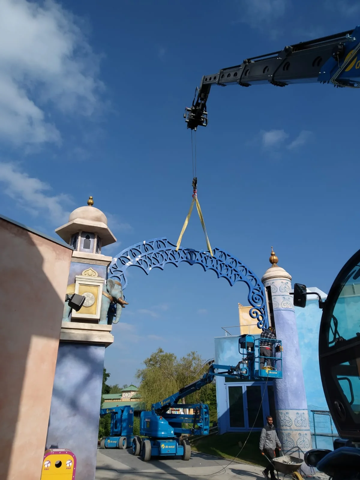 Crane installation of the entrance portal to the Bollywood area: a large blue openwork arch, fretted like Indian goldsmithery, lifted by a yellow crane between two sand-coloured Mughal columns with gilded domes; clear sky behind, build-site tools visible at the bottom
