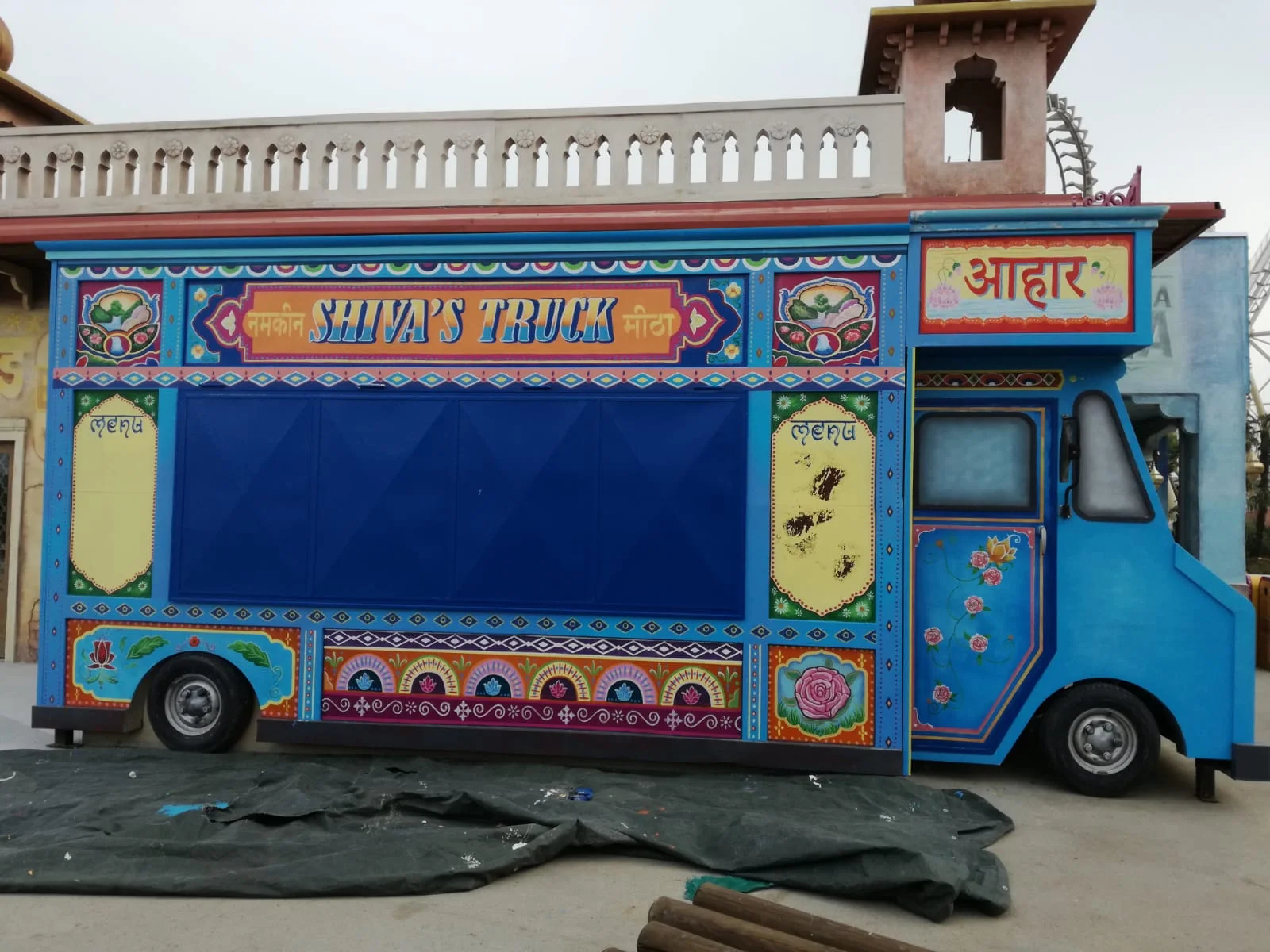 Side view of the completed Shiva's Truck: a wide side panel with a blue frame, Hindi lettering and 'SHIVA'S TRUCK' across a red central band, decorative rosettes, painted flowers, gilded edges, Mughal geometries, black wheels; above it the pink-magenta temple-style crenellation, with the park's architecture and coaster in the background