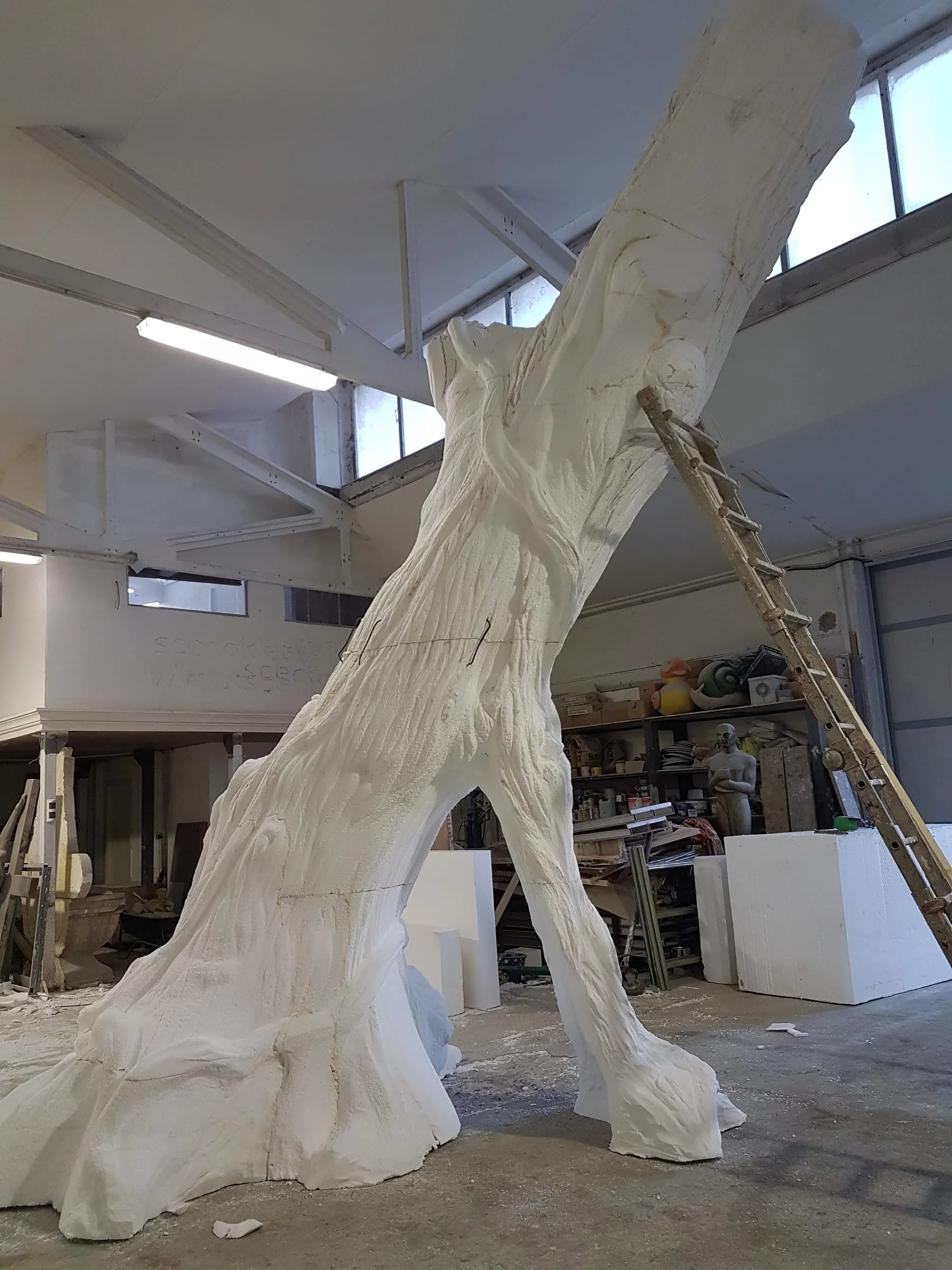 A second giant tree trunk in white polystyrene, with a three-branch fork, mounted in the workshop under the skylights. Beside it a ladder leaning against the trunk: the piece is several metres tall.