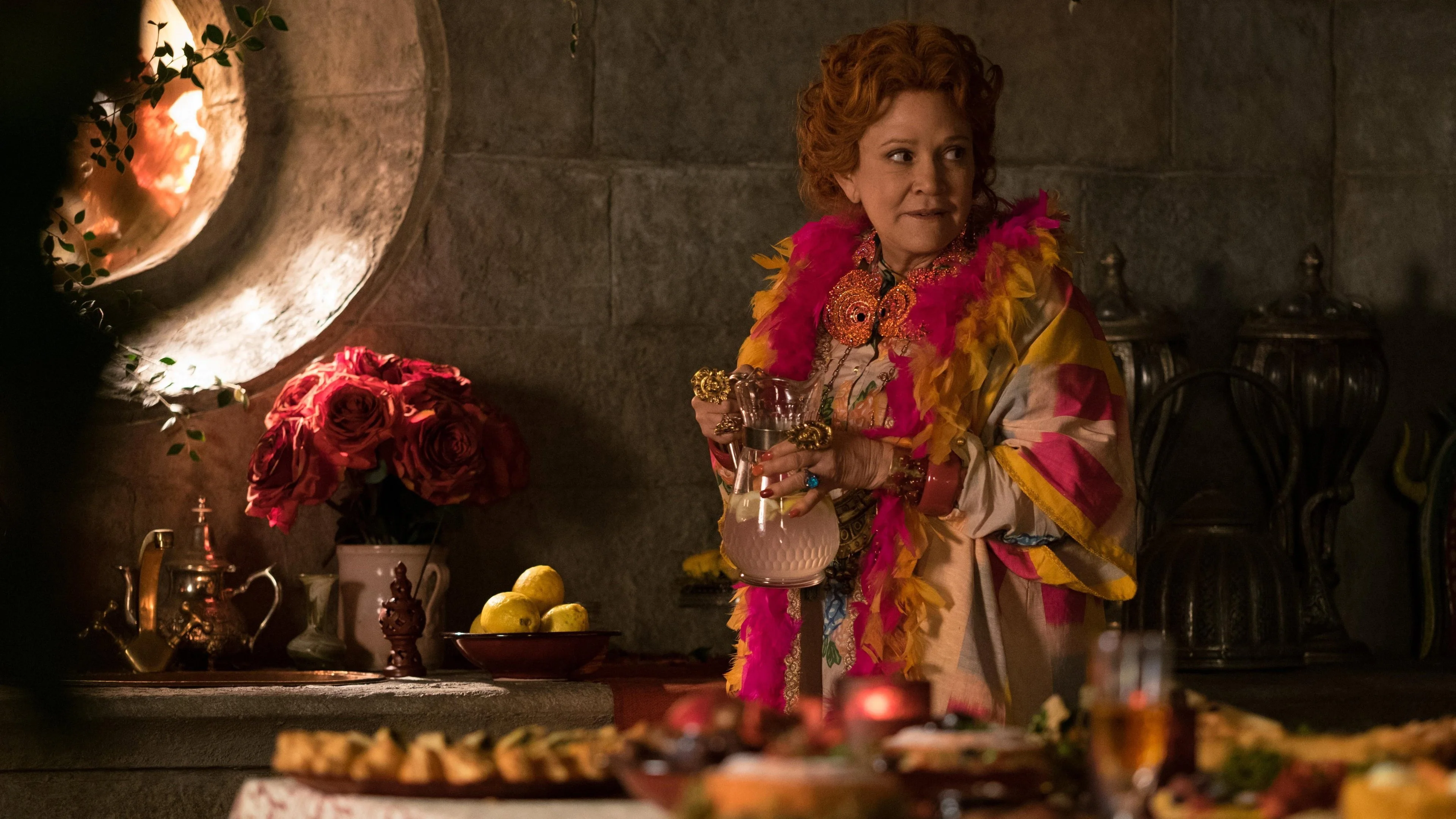 Carrie Fisher as elderly Hazel — red hair, fuchsia scarf — pours herself a drink next to a large circular ogival portal lit by fire. Red roses and crockery on the table.