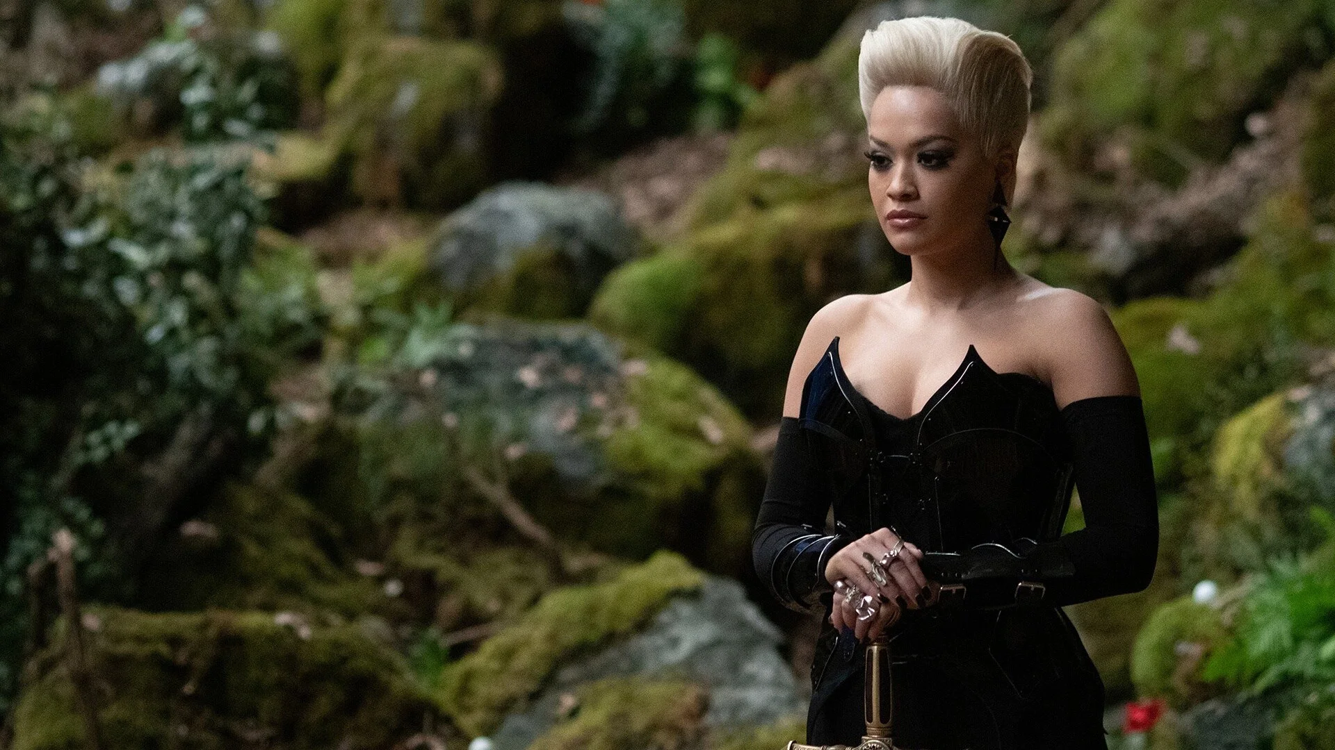 Rita Ora as young Hazel — platinum blonde hair, black corset and gloves — standing in the forest among moss-covered rocks and ferns. The stone is the one we sculpted.