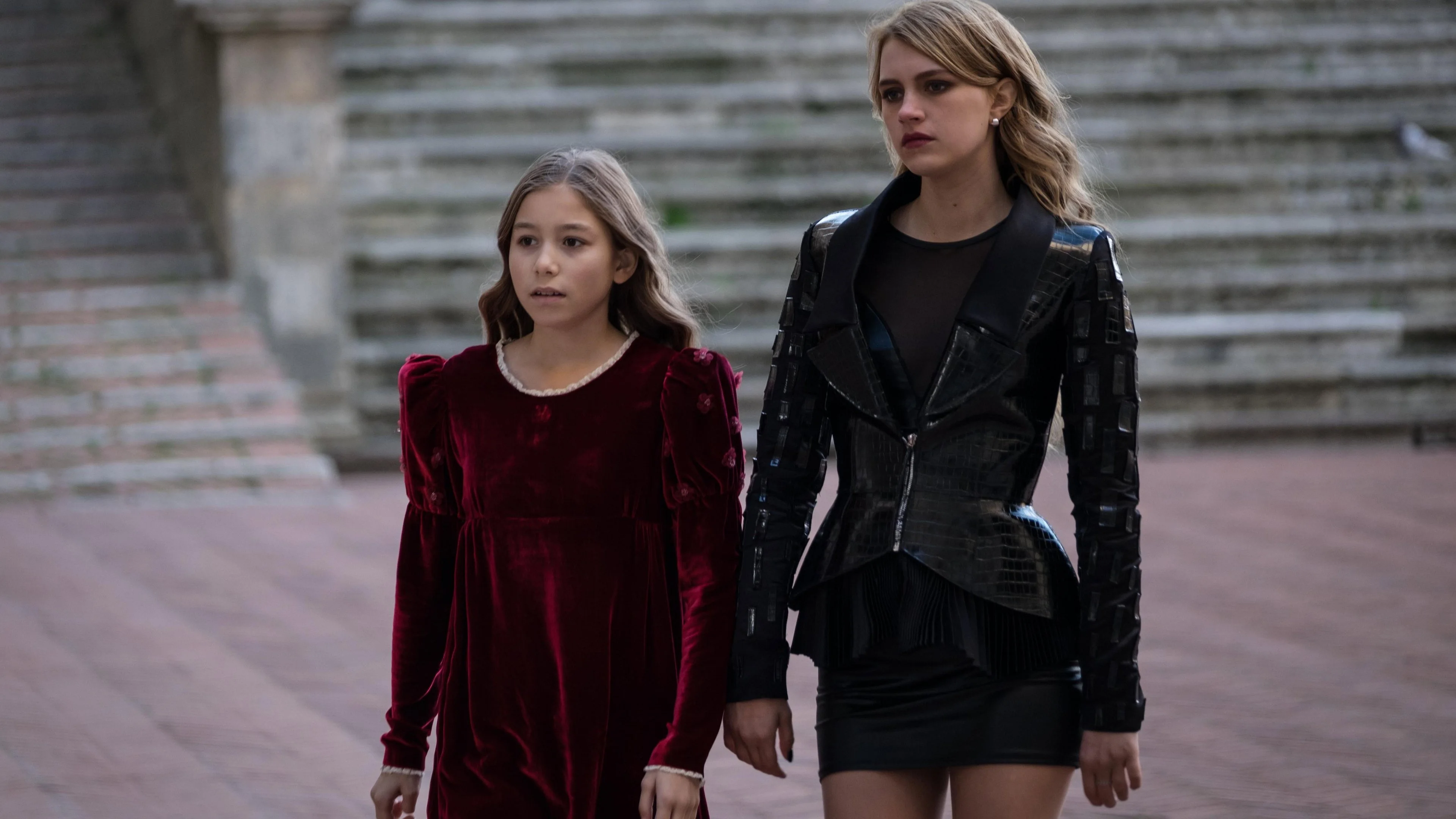Violet (Kiera Milward) in a burgundy velvet dress walks beside young Hazel (Rita Ora) — black leather jacket, short skirt — across a monumental square of the Etruscan village, grey stone staircase behind.