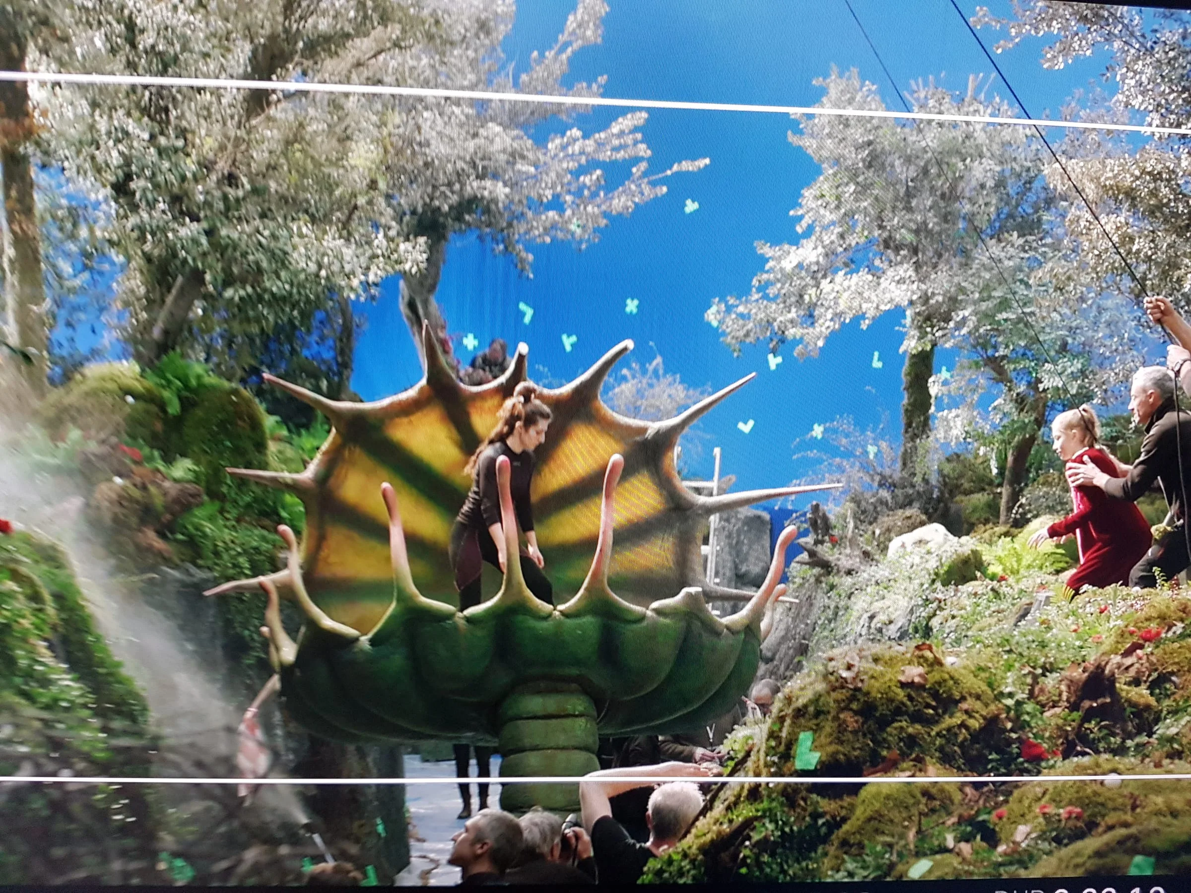 Film shoot in progress: a carousel in the shape of a horned yellow and green spiny-shell creature, with a girl on top. Below, crew and technicians. In the background a blue cyclorama with butterflies.