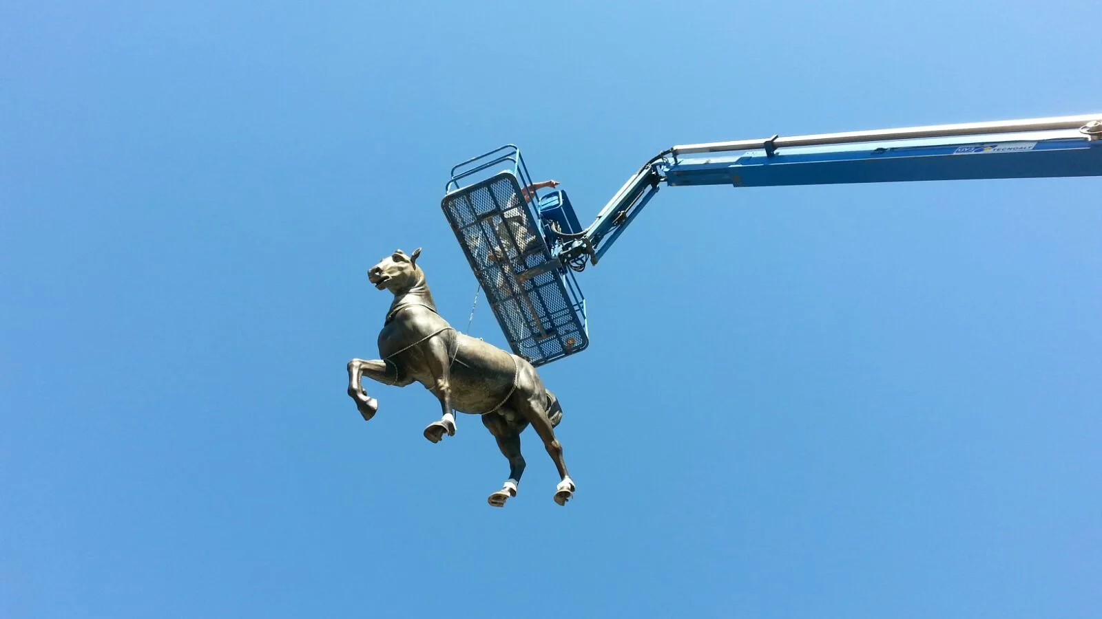 Una delle statue equestri in finto bronzo sollevata in aria da un cestello di una gru telescopica contro un cielo azzurro: la sagoma è quella della quadriga di San Marco, sospesa a mezz'aria sopra il piazzale di Cinecittà