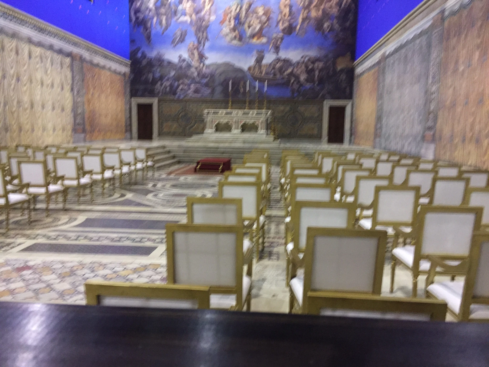 Vista d'insieme della cappella papale ricostruita: file di sedie d'oro, pavimentazione cosmatesca a mosaico geometrico, balaustra centrale, altare con candelabri, riproduzione del Giudizio Universale sopra il fondale, cyclorama blu sullo sfondo