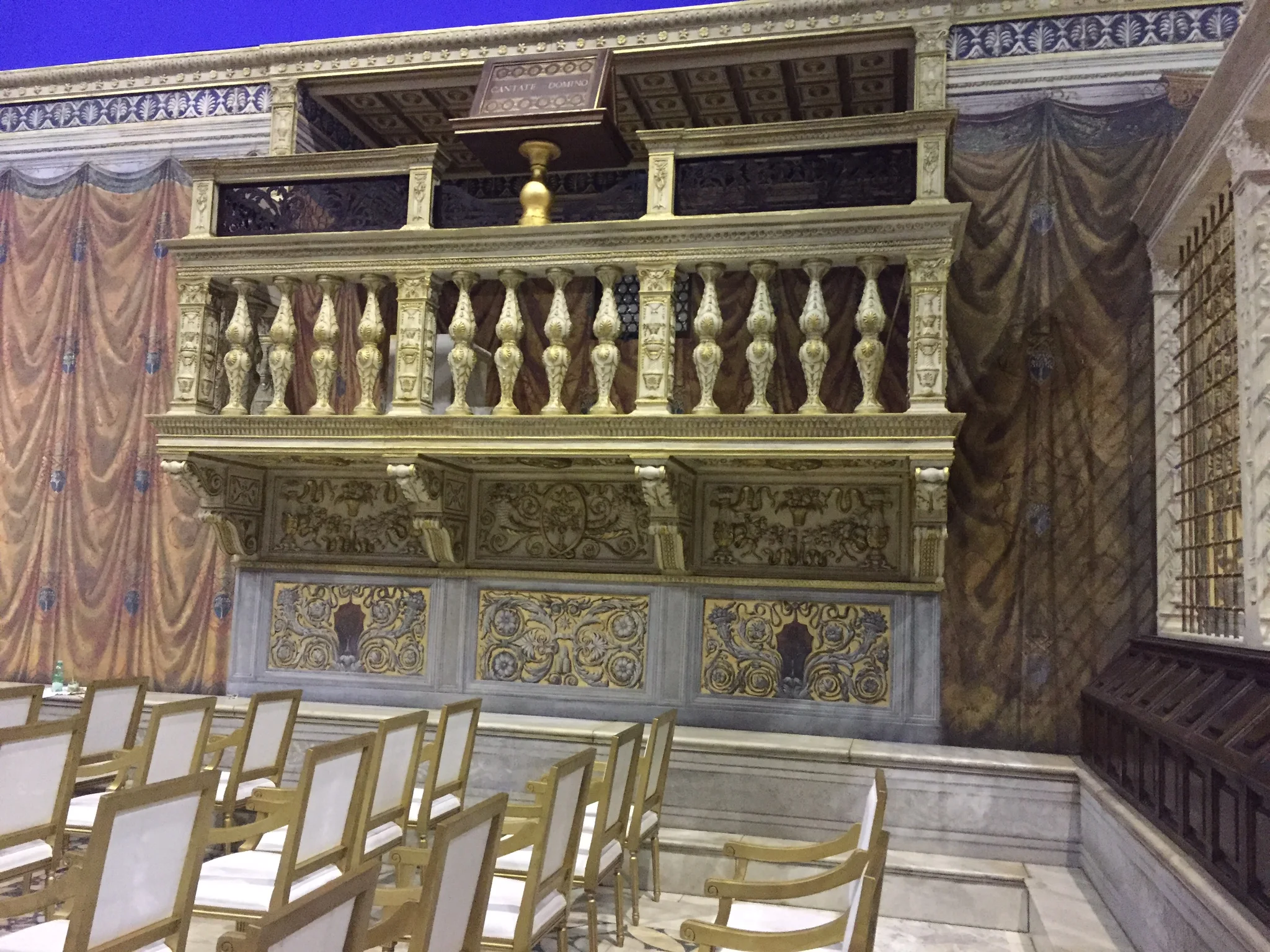 Loggia delle Benedizioni ricostruita: balaustra in finto marmo dorata, colonne tornite, pannelli a bassorilievo sotto, tendaggi di velluto rosso ai lati, vista parziale delle sedie di velluto in primo piano